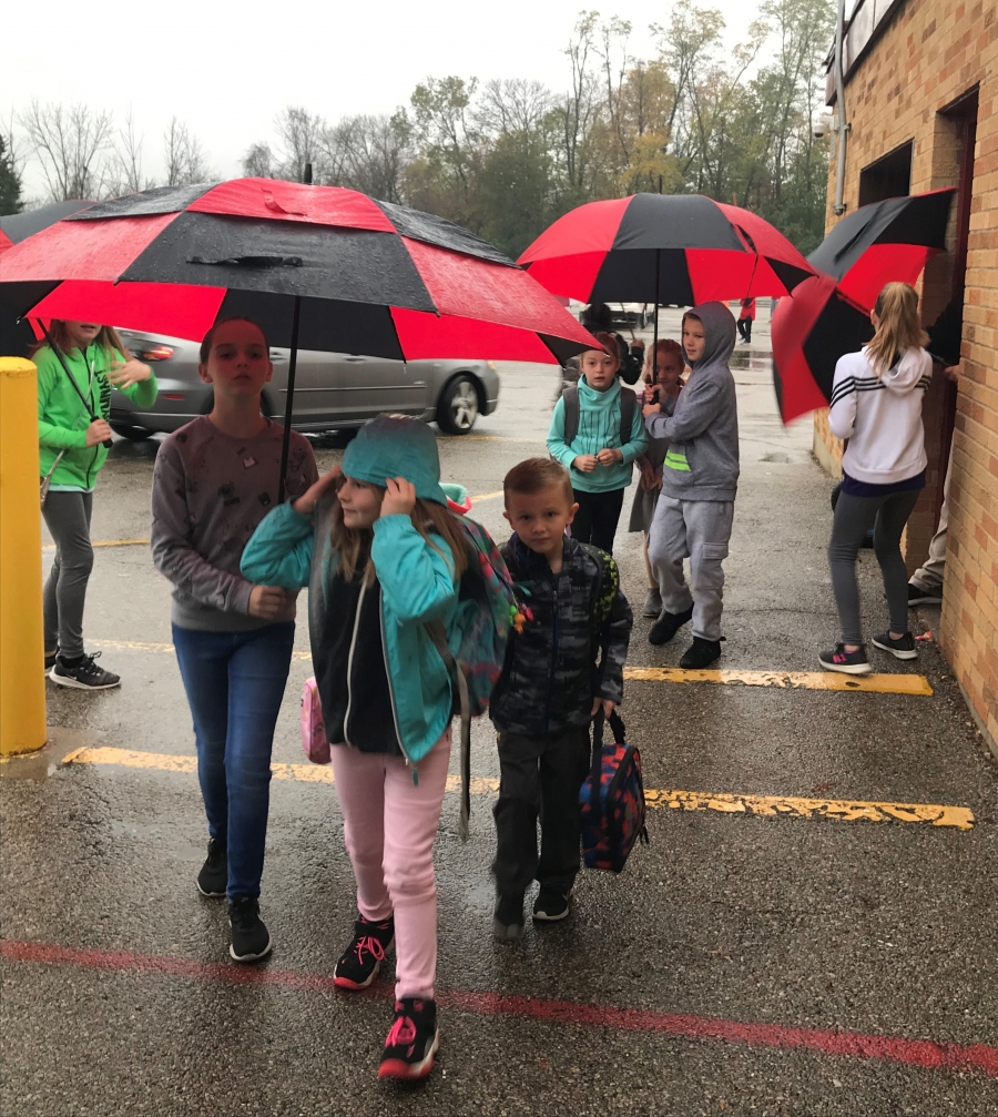 Fifth graders with umbrellas walk younger students in from carline on rainy days.